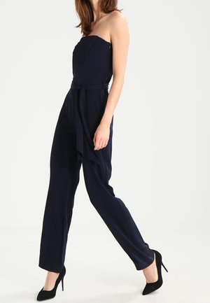 Marineblauwe strapless jumpsuit met een aansluitende bodice, wijde pijpen en een gestrikte riemdetail. Draag het met zwarte stilettohakken op een effen achtergrond.