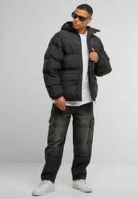 Giacca a puffo nera con cappuccio, dotata di zip frontale e due tasche frontali. Indossata con pantaloni cargo grigi e sneakers bianche.