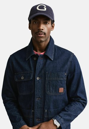 Uomo con cappellino da baseball blu navy con logo "G", giacca di denim scuro con tasche frontali e orologio da polso d'argento su sfondo bianco.