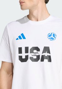 Bijela pamučna majica sa plavim Adidas logotipom, crnim tekstom "USA" s gradijent efektom i plavim kružnim grafičkim motivom na lijevoj strani prsnog dijela.
