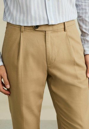 Man wearing light brown pleated trousers and a tucked-in long-sleeve striped shirt, standing with hands relaxed at sides.