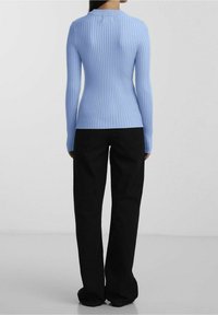 Frau steht mit dem Rücken zur Kamera, trägt einen hellblauen gerippten Langarmpullover und lockere schwarze Hosen vor einem schlichten Hintergrund.