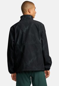 Schwarze Jacke mit Wellenmuster, hohem Kragen, elastischen Bündchen und lockerer Passform. Der Stoff wirkt glatt und leicht.