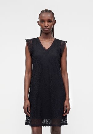 Žena s vrkočmi vo vlasoch, ktorá má na sebe čierne čipkované bezrukávové šaty s výstrihom do V a vlnkovanými okrajmi, stojaca pred jednoduchým svetlým pozadím.