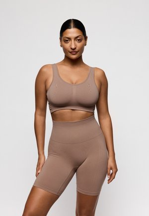 Vrouw met donker haar in een strakke knot, gekleed in een bijpassende bruine sportbeha en hoge taille fietsbroek, staand tegen een effen lichte achtergrond.