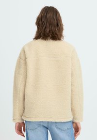 Beżowa kurtka polarowa o teksturowanej, puszystej powierzchni, luźnym kroju i opadających ramionach. Posiada prosty dół oraz brak widocznych zapięć.