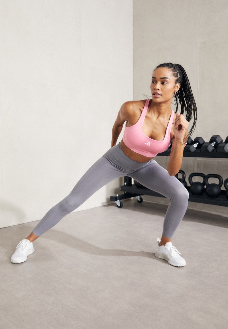 Rosa Sport-BH, graue Leggings und weiße Sneakers. Das Modell befindet sich in einer Ausfallschritt-Position und präsentiert gut sitzende Sportbekleidung vor einem neutralen Hintergrund.