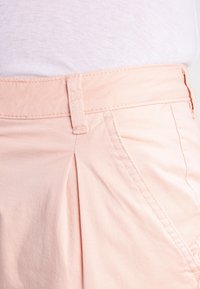 Světle růžové kalhoty s hladkou texturou látky, vybavené pasem a řaseným designem. Podél švů jsou viditelné stehy.