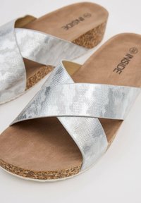 Sandalias de tiras cruzadas plateadas con parte superior metálica texturizada, suela de corcho y plantilla marrón suave. Diseño liviano para uso informal.