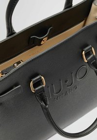 Borsa a tracolla in pelle nera con superficie testurizzata, dettagli in hardware dorato, doppi manici e logo impresso sul davanti. L'interno è di un beige chiaro.