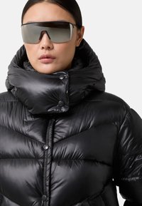 Zwarte pufferjas met een hoge kraag, glossy textuur en knoopaccenten. Model draagt een oversized donkere zonnebril.