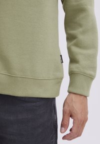 Sudadera verde oliva con puños y dobladillo acanalados, que presenta una pequeña etiqueta negra en el lateral. El material parece suave y texturizado.