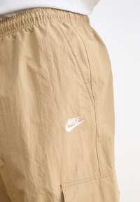 Beige cargo shorts lavet af letvægtsstof, med en elastisk talje og et hvidt Nike-logo på siden.