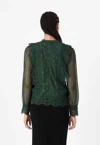 Blusa di pizzo verde con maniche lunghe trasparenti, spalle arruffate e un bordo a scallop intricatamente lavorato. Delicati motivi floreali su tutto il capo.
