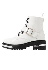 Bottine en cuir verni blanc à texture crocodile, lacets noirs, boucle en métal, semelle épaisse noire et accent argenté sur le talon.