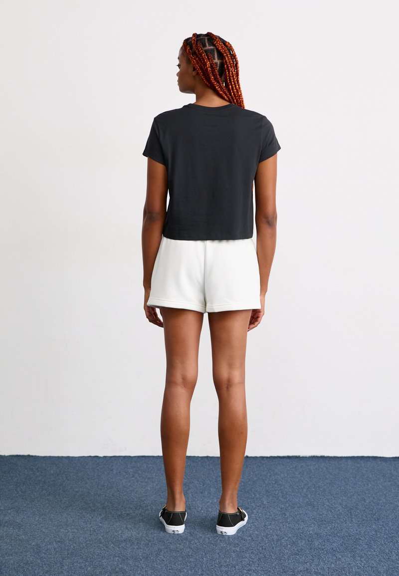 Camiseta negra corta de manga corta, combinada con pantalones cortos blancos. El modelo lleva zapatillas negras. El fondo es claro y sencillo.