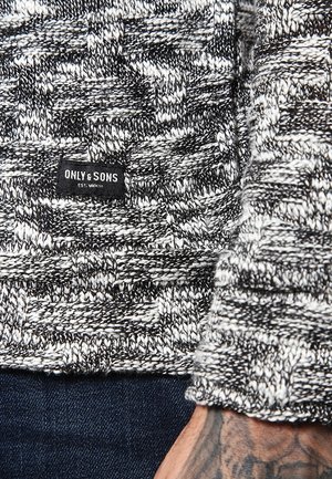 Schwarz-weißer Strickpullover mit gewebter Struktur, mit dem Etikett "ONLY & SONS", getragen über dunkelblauen Jeans, mit einem tätowierten Unterarm zu sehen.