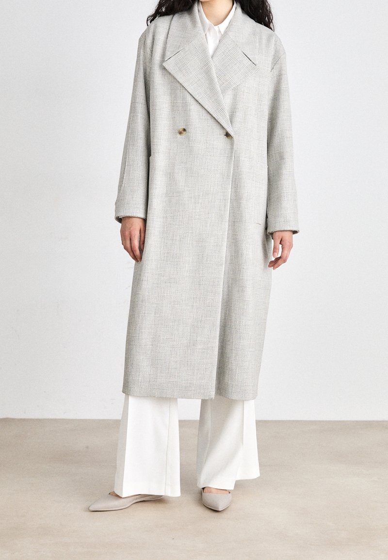 Lichtgrijze geruite jas met een dubbelzijdig ontwerp, grote revers en lange mouwen, gedragen over witte wijde broeken en platte schoenen.