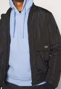 Giacca bomber nera in nylon con chiusura a zip, tasche frontali e tasca sulla spalla, indossata sopra un felpa con cappuccio blu chiaro.