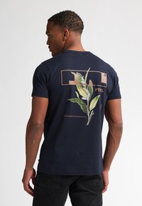 Navyblauwe katoenen t-shirt met een bloemontwerp op de rug, met groene bladeren en witte bloemen binnen een rechthoekige omlijning.