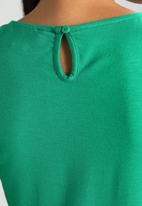 Maglietta verde con una texture morbida, caratterizzata da un'apertura a chiave sul retro fissata da un bottone rotondo. Design semplice e informale.