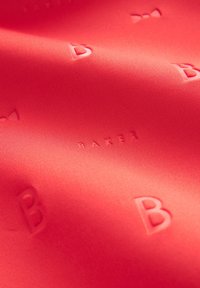 Un tessuto di colore rosso corallo presenta lettere embossate 'B' e un motivo a fiocco, mostrando una texture liscia e un design elegante e moderno.