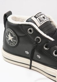 Czarne skórzane sneakersy Converse All Star w wersji wysokiej z białym futerkiem, srebrnymi oczkami, czarnymi sznurowadłami i okrągłą naszywką z logo.