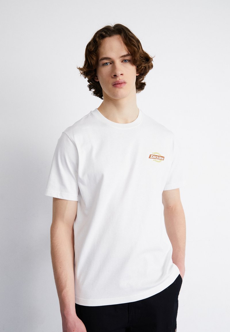 Camiseta de algodón blanca con un pequeño logo "Dickies" en naranja y amarillo en el lado izquierdo del pecho, cuello redondo y mangas cortas.