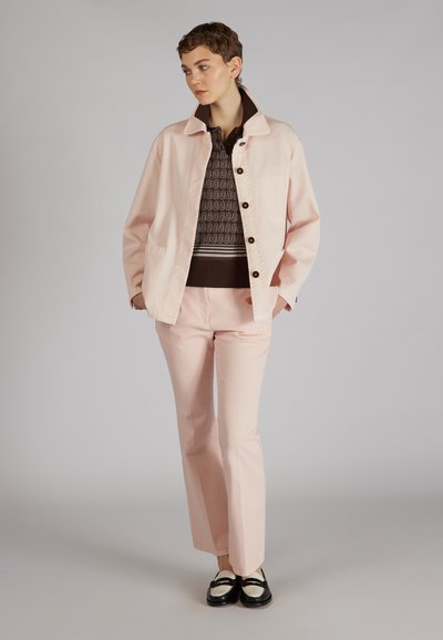 Chaqueta de lona rosa clara con cuello, cierre de botones, combinada con pantalones de campana rosas a juego y un suéter marrón con patrón por debajo. Zapatos negros.