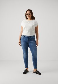 White, short-sleeve cotton top with lace detailing, paired with blue, fitted denim jeans and black loafers, against a neutral background.