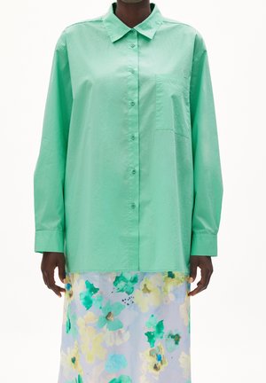 Person trägt ein übergroßes, mintgrünes Hemd und einen pastellfarbenen, geblümten Rock mit grünen, gelben und blauen Blumen im Aquarellstil.