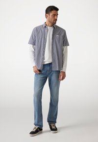 Camisa de manga corta a rayas en azul marino y blanco, con cuello abotonado y un bolsillo, combinada con jeans azul claro y zapatillas negras.