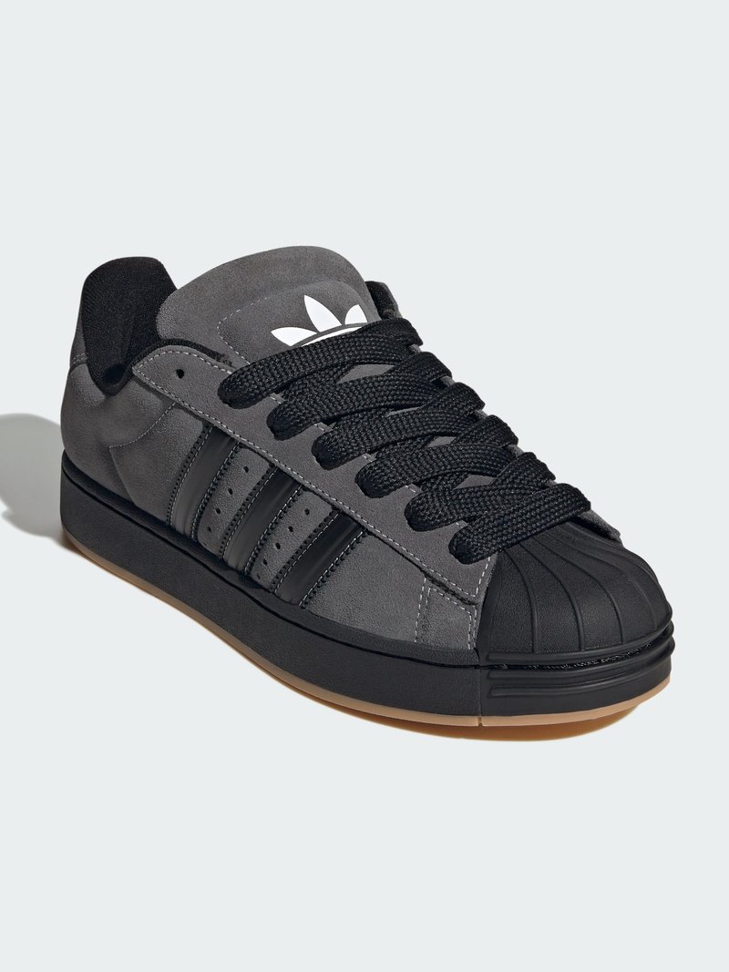 Grå mockasinsneaker med svart gummiskydd på tå, tre svarta ränder och svarta skosnören. Har vit logga på tungan och gummisula.