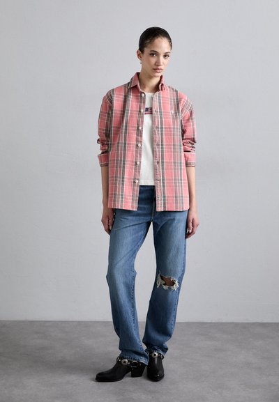 Camisa de cuadros rosa sobre una camiseta blanca, combinada con jeans azules rasgados y zuecos negros de tacón, con un fondo neutral.