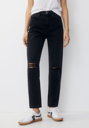 Personne portant un jean noir déchiré taille haute et des baskets blanches avec des rayures noires, debout sur un fond blanc uni.