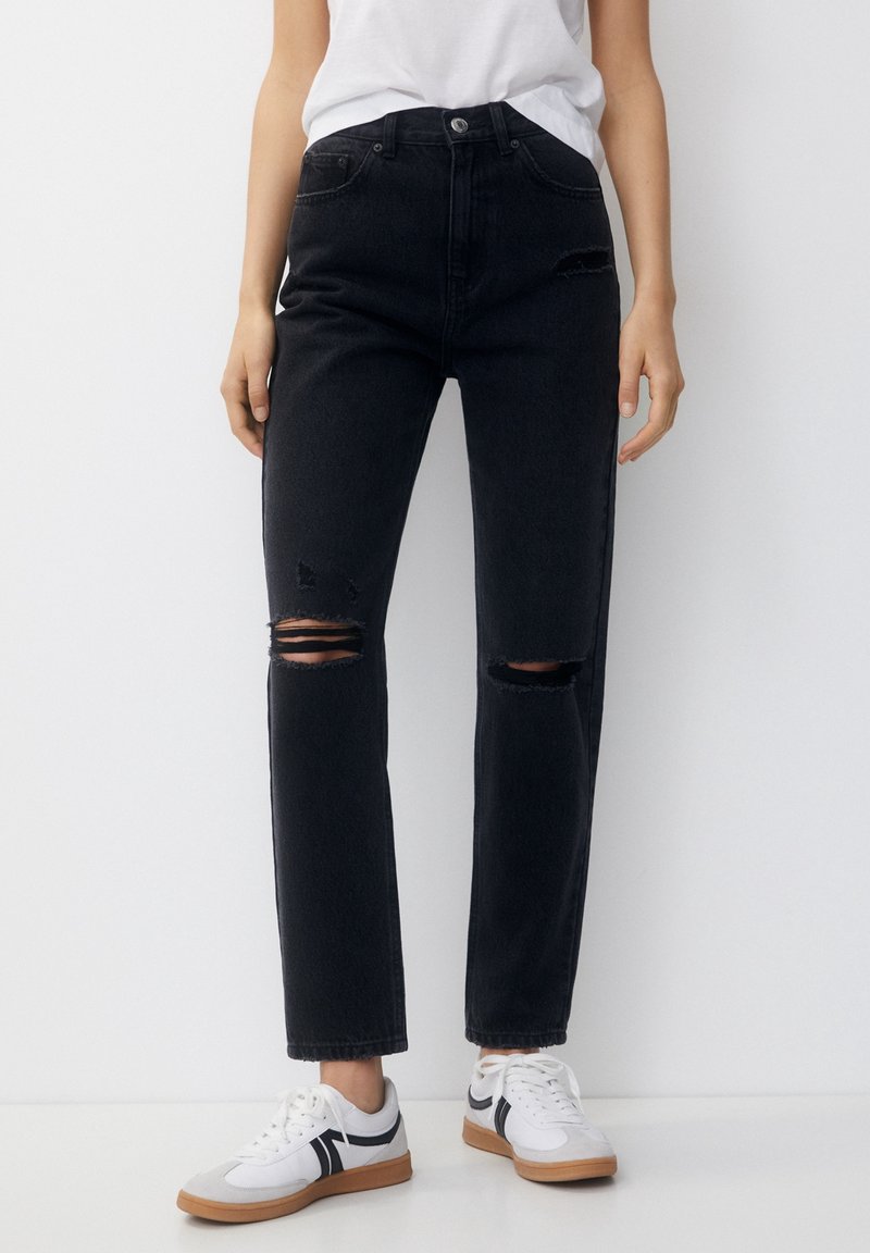 Personne portant un jean noir déchiré taille haute et des baskets blanches avec des rayures noires, debout sur un fond blanc uni.