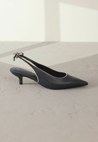 Mustekka nahkainen slingback-korkokenkä, jossa on terävä varpaan muoto, kontrastiväriset vaaleat reunat, hoikka keskipitkä korko ja koristeellinen solmukuvio takana.