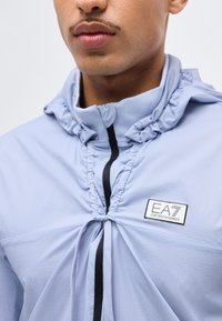 Κοντινή λήψη του κάτω μέρους του προσώπου και του λαιμού ενός ατόμου που φοράει ένα ανοιχτό μπλε μπουφάν με φερμουάρ και σούρες στο γιακά, με το λογότυπο EA7 Emporio Armani στο στήθος.