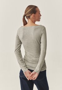 Graues Langarmoberteil mit weitem Bootsausschnitt, taillierter Passform und geraffter Textur an den Seiten, kombiniert mit dunklen Jeans.