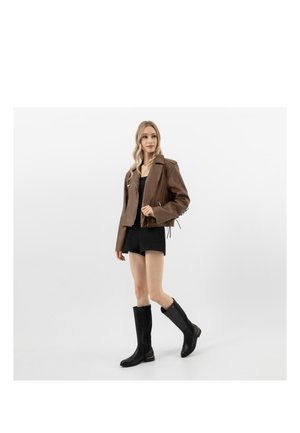 Žena v hnědé kožené bundě, černých šortkách a černých kozačkách po kolena stojí před jednoduchým bílým pozadím.