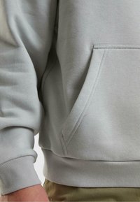 Ljusgrå sweatshirt med detalj på ficka och ärm på en person som bär khakifärgade byxor, som visar tygtextur och sömmar.