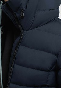 Marineblauwe pufferjas met gewatteerd ontwerp, hoge kraag, ritssluiting aan de voorkant en een zwart trekkoord bij de halslijn. Zacht, textuurrijk materiaal.