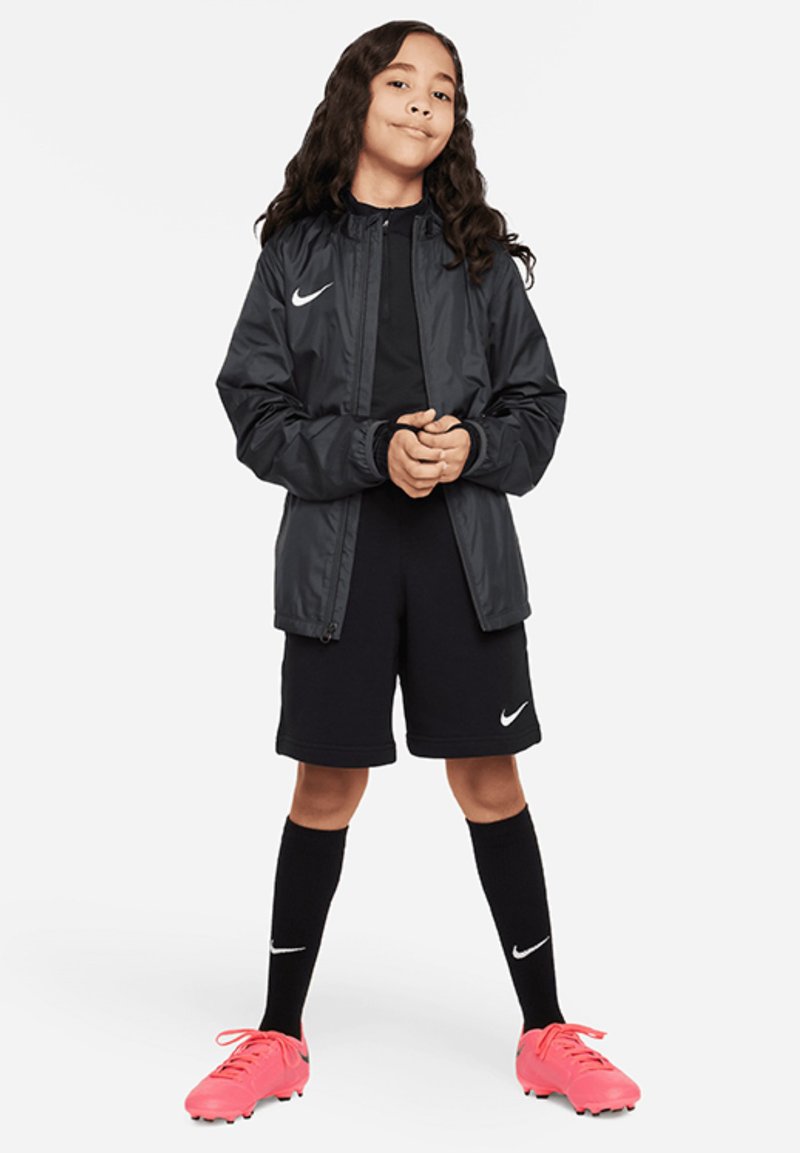 Rompevientos negro de Nike sobre una camiseta y pantalones cortos negros. Calcetines negros con acentos blancos y zapatillas de fútbol rosas brillantes completan el look atlético.