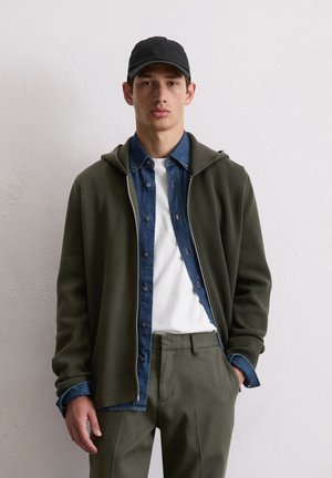 Green zip-up hoodie layered over a blue denim shirt, white T-shirt underneath, and olive green pants. Black cap completes the outfit.
