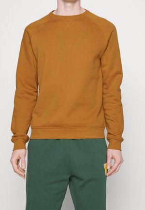 Mann trägt einen langärmeligen Sweatshirt in Senfgelb und dunkelgrüne Hose mit einem kleinen gelben Fleck am linken Oberschenkel.