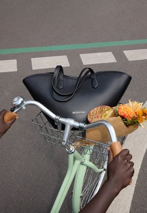 Les mains d'une personne sur le guidon d'un vélo avec un sac Valentino noir et un bouquet de fleurs dans le panier avant, roulant sur une route marquée.