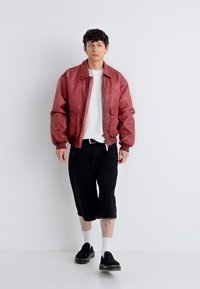 Νέος άνδρας περπατάει δίπλα σε λευκό τοίχο φορώντας κόκκινο μπουφάν τύπου bomber, άσπρο μπλουζάκι, μαύρο σορτς μέχρι τα γόνατα, άσπρες κάλτσες και μαύρα παπούτσια χωρίς κορδόνια.