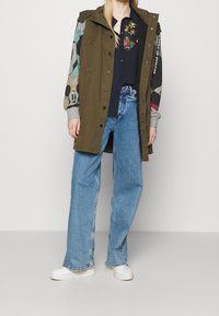 Cappotto lungo verde oliva con maniche a fantasia colorate, camicia blu scuro e jeans larghi azzurri. Sneakers bianche completano il look.