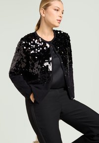 Cardigan nero con paillettes sul davanti, scollatura rotonda e maniche lunghe. Indossato sopra una maglia nera, abbinato a pantaloni neri.