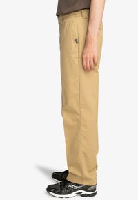 Light khaki pants with a straight leg design, made from cotton, feature belt loops and side pockets. Worn with black athletic sneakers.